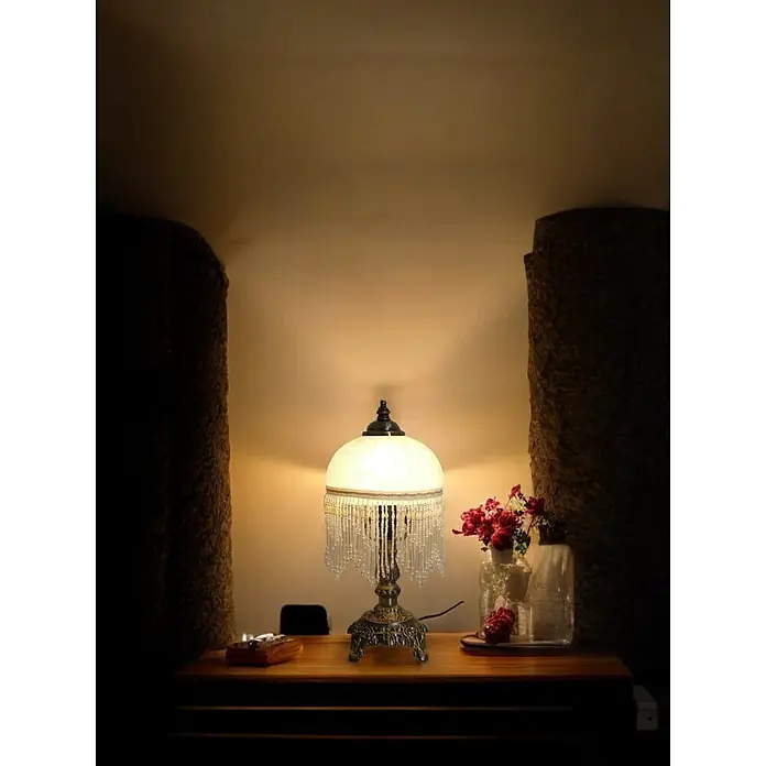 Tischleuchte Weide Landhaus Stil messing -  - Verzierte Tischlampe aus Bronze mit weißem Glasschirm und Perlenfransen, die einen Holztisch beleuchtet.