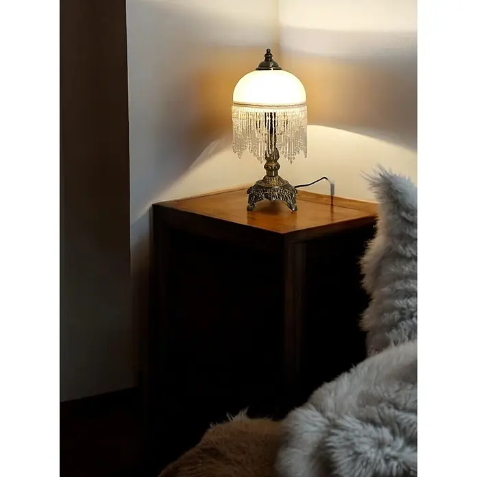 Tischleuchte Weide Landhaus Stil messing -  - Eine beleuchtete Vintage-Tischlampe mit Bronzefuß und Perlenfransen-Schirm steht auf einem Holznachttisch.