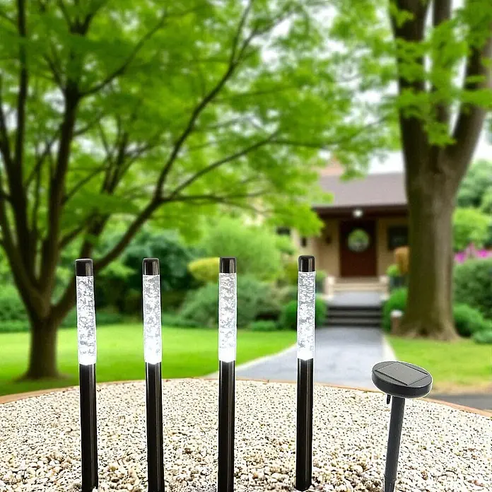 Solarleuchte Bubble 4er Set LED-Kaltweiß -  - Vier beleuchtete Solar-Gartenleuchten mit Blaseneffekt und ein Solarpanel stehen in einem Gartenbeet aus Kieselsteinen.