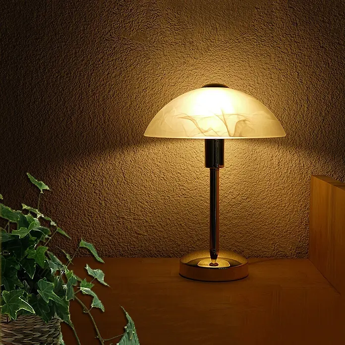 Tischleuchte Mischa Messing E14 -  - Beleuchtete Tischlampe mit gewölbtem, mattiertem Glasschirm und Metallsockel, die warmes Licht auf eine strukturierte Wand wirft.