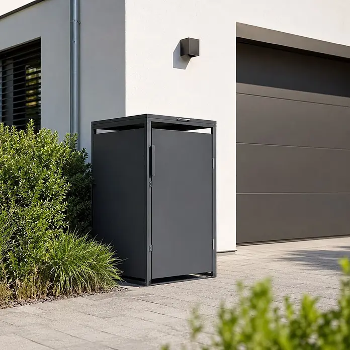Norlinio Mülltonnenbox Vario, Anthrazit, für 1 Tonne -  - Dunkelgrauer Metall-Außenschrank neben Haus und Garage, auf gepflastertem Bereich mit Sträuchern.