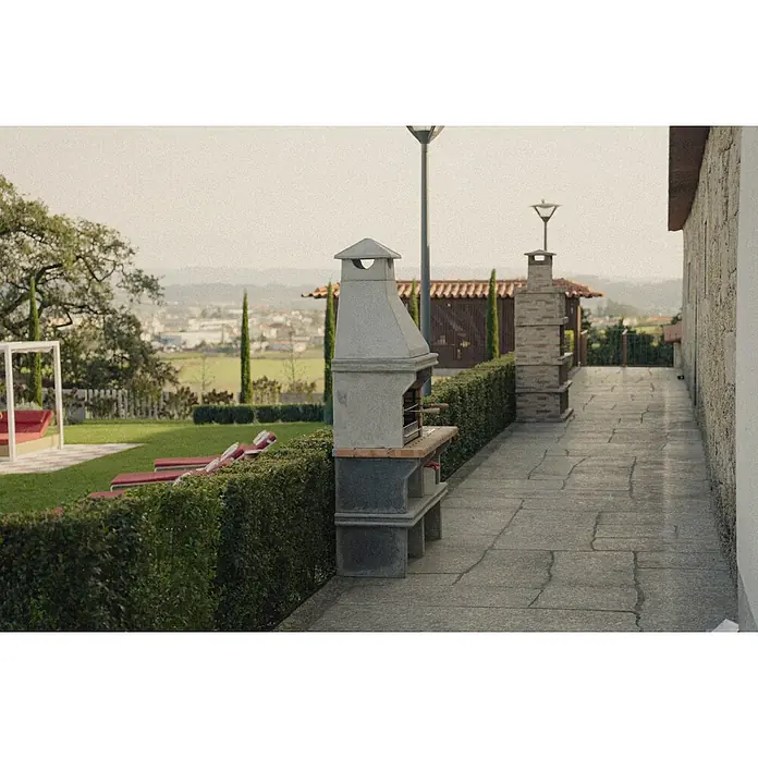 Sotnas Barbacoa de obra Ibiza - Gris/Ladrillo, An x Pr x Al: 168 x 60 x 219 cm, Superficie parrilla: 60 x 38 cm, Con fregadero - Una barbacoa de piedra con chimenea se encuentra en un patio pavimentado junto a un seto verde, con vistas a un jardín y una ciudad lejana.