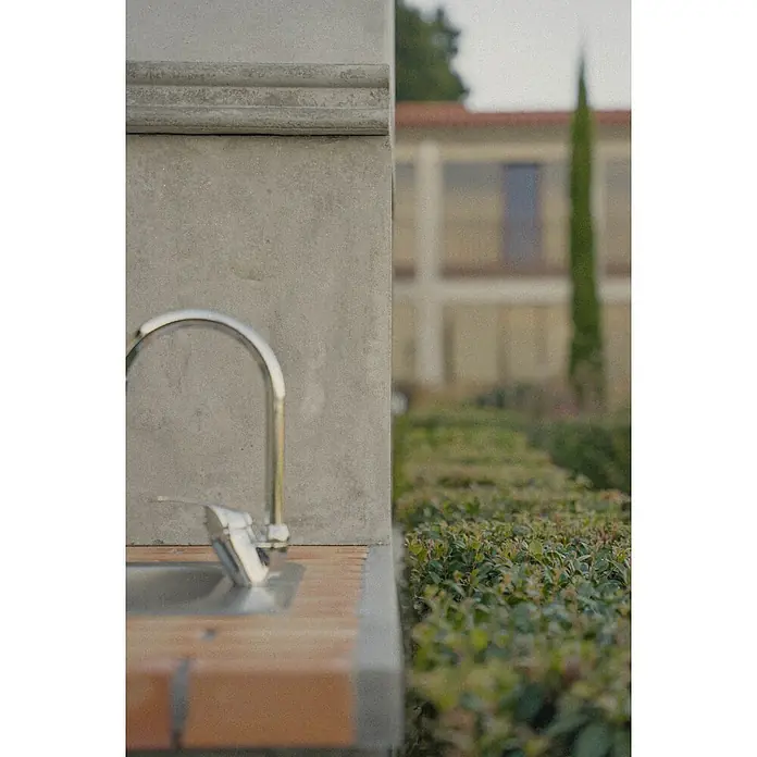 Sotnas Barbacoa de obra Ibiza - Gris/Ladrillo, An x Pr x Al: 168 x 60 x 219 cm, Superficie parrilla: 60 x 38 cm, Con fregadero - Primer plano de un grifo exterior metálico sobre una estructura de hormigón y ladrillo, con un jardín borroso al fondo.