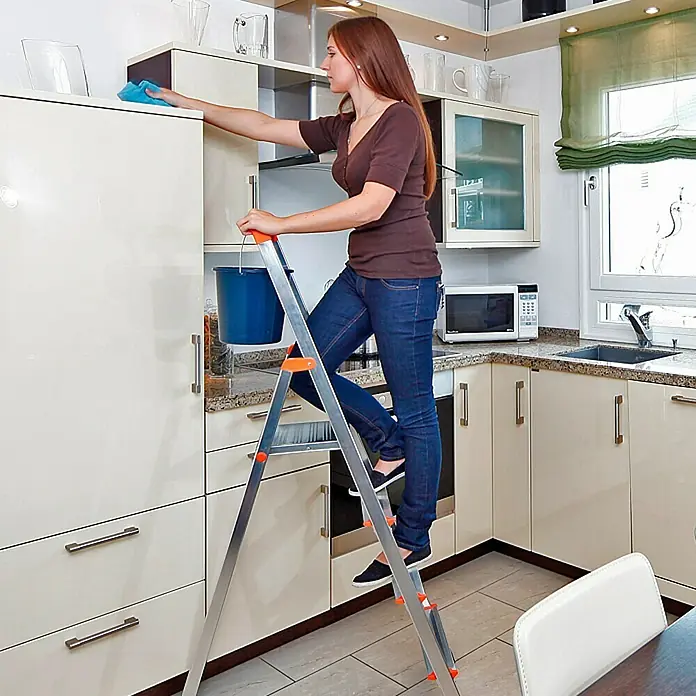 Krause Monto Escalera de tijera - Altura de trabajo: 2,85 m, Número de peldaños: 4 ud., Carga soportada: 150 kg, Aluminio - Mujer limpiando armario de cocina con taburete. El taburete es de aluminio con detalles naranjas, ideal para uso doméstico.