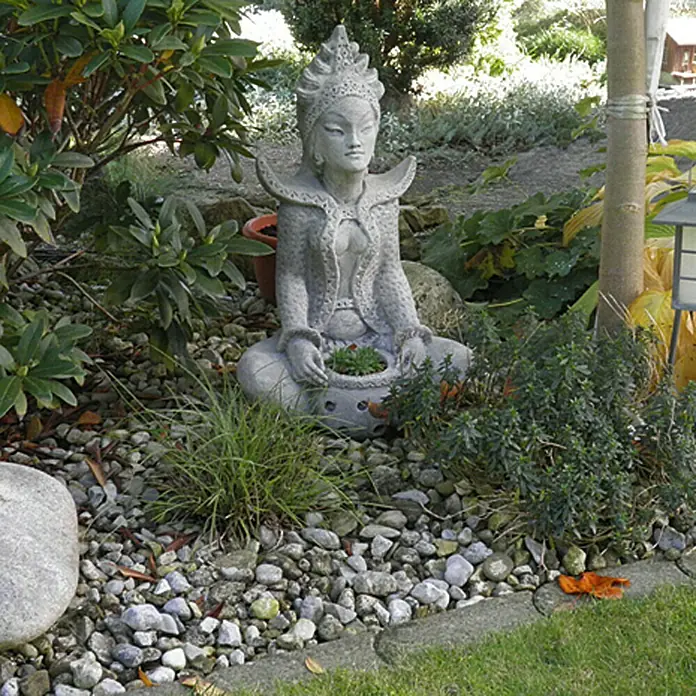 Min2C Flusskiesel - Bunt, Körnung: 16 mm - 32 mm, 25 kg - Eine Steinbuddhafigur sitzt inmitten eines Gartens, umgeben von Kies und Pflanzen, die eine friedliche und besinnliche Atmosphäre ausstrahlen.
