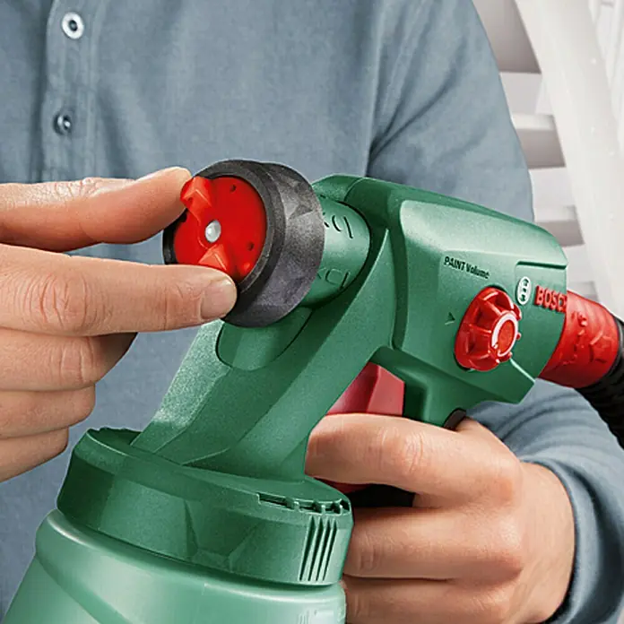 Bosch Sistema de pulverización de pintura PFS 2000 - Apto para: Todos los colores, 440 W, Caudal: 0 l/min - 0,2 l/min, Velocidad de trabajo: 1,5 m²/min - Primer plano de una pistola de pintura Bosch sostenida por una persona, enfocándose en la boquilla y el ajuste del volumen de pintura.