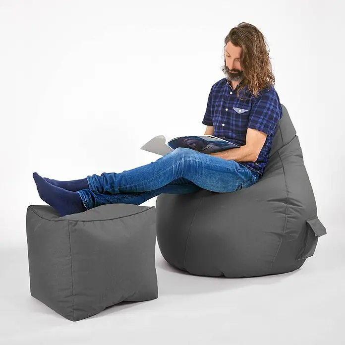 Green Bean Sitzhocker Cube mit EPS Füllung 40x40x40 cm Grau -  - Mann entspannt auf grauem Stoff-Sitzsack mit Füßen auf passendem Hocker, liest Magazin.