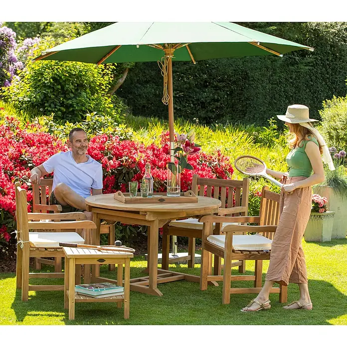 Teak Beistelltisch Cuneo 2 Ebenen 50 x 50 cm Massivholz wetterfest und edel -  - Eine Holzgartenmöbelgarnitur mit rundem Tisch und Stühlen, die von zwei Personen in einem üppigen Garten genutzt wird.