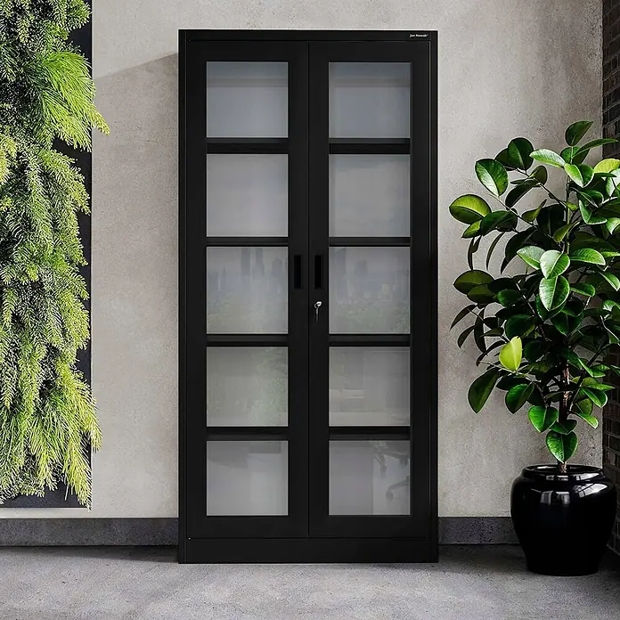 Jan Nowak Vitrinenschrank AMELIA, 90x185x40 cm, Schwarz -  - Schwarzer Metallschrank mit zwei mehrfeldrigen Glastüren und Schloss, steht an einer Betonwand zwischen grünen Pflanzen.