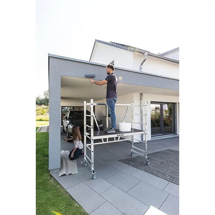 ML Klappgerüst & Teleskopausleger 150x60cm Arbeitshöhe  3m Traglast 150kg -  - Mobiles Aluminium-Rollgerüst mit Plattform, von einem Mann zum Streichen einer Hausfassade genutzt. Mit Rollen für leichte Bewegung.