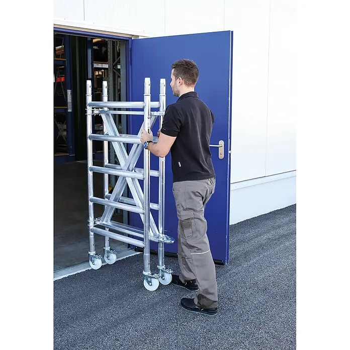 Klappgerüst Plattformgröße 180x135cm Arbeitshöhe bis 3m inkl. 4x Lenkrollen -  - Person schiebt ein kompaktes Aluminium-Fahrgerüst auf Rollen durch eine Tür, was den einfachen Transport hervorhebt.