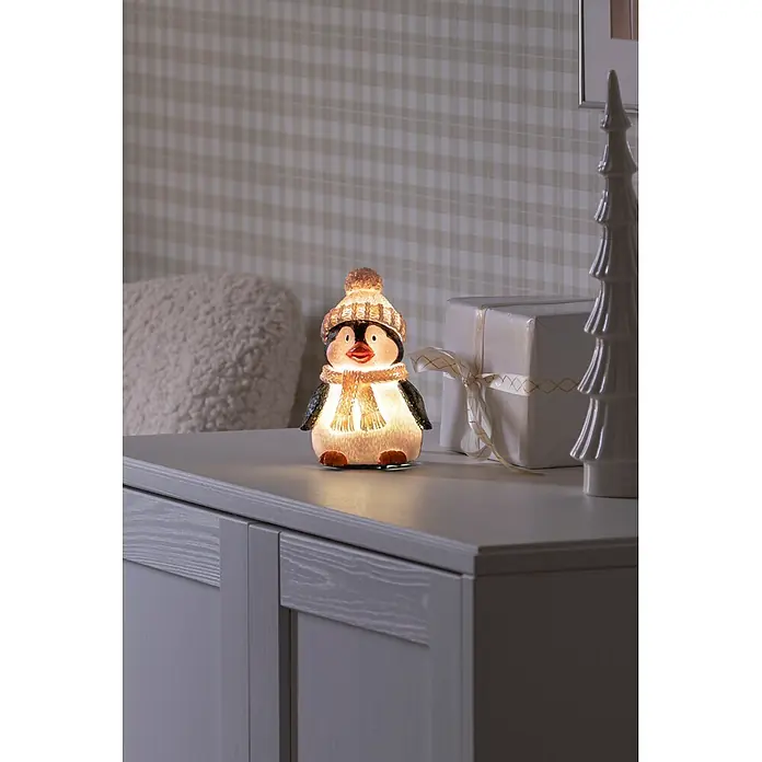 Konstsmide LED-Weihnachtsdekofigur - B x H: 9,5 x 15,5 cm, Lichtfarbe: RGB, Batteriebetrieben, LED - Leuchtender Dekopinguin mit Mütze und Schal auf einem grauen Schrank, spendet warmes Licht.