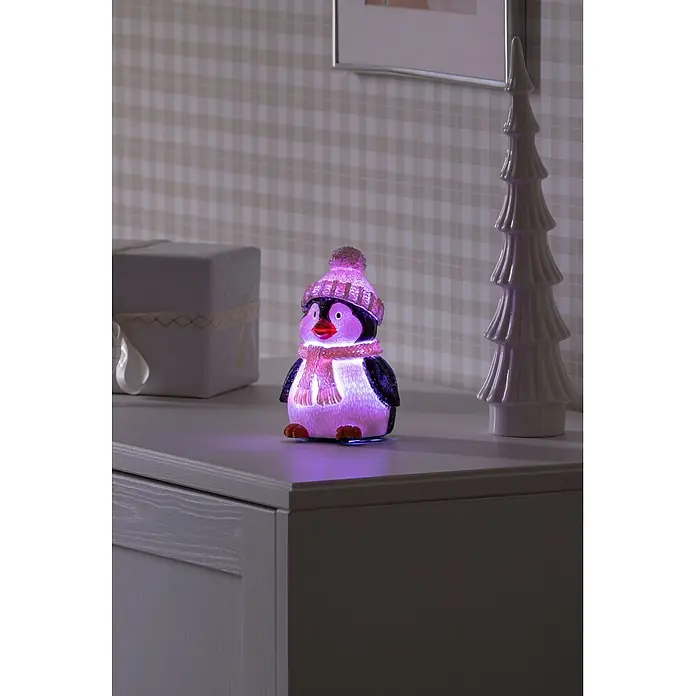 Konstsmide LED-Weihnachtsdekofigur - B x H: 9,5 x 15,5 cm, Lichtfarbe: RGB, Batteriebetrieben, LED - Eine leuchtende Pinguinfigur mit rosa Mütze und Schal sitzt auf einer grauen Kommode und strahlt lila Licht aus.