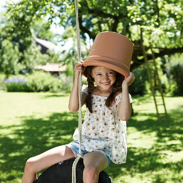 Elho Green Basics Maceta para planta - Diámetro exterior máx.: 11 cm, Plástico, Tono rojo - Una niña alegre en un columpio de neumáticos lleva una maceta como sombrero, capturando un momento lúdico e imaginativo al aire libre.