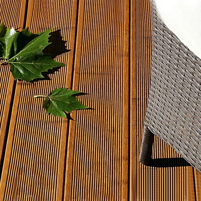 CoBaM Terrassendiele - Bambus, 220 x 14 x 2 cm, Braun - Detailaufnahme eines braunen Holzdecks mit grünen Blättern und einem Teil eines grauen Korbsessels.