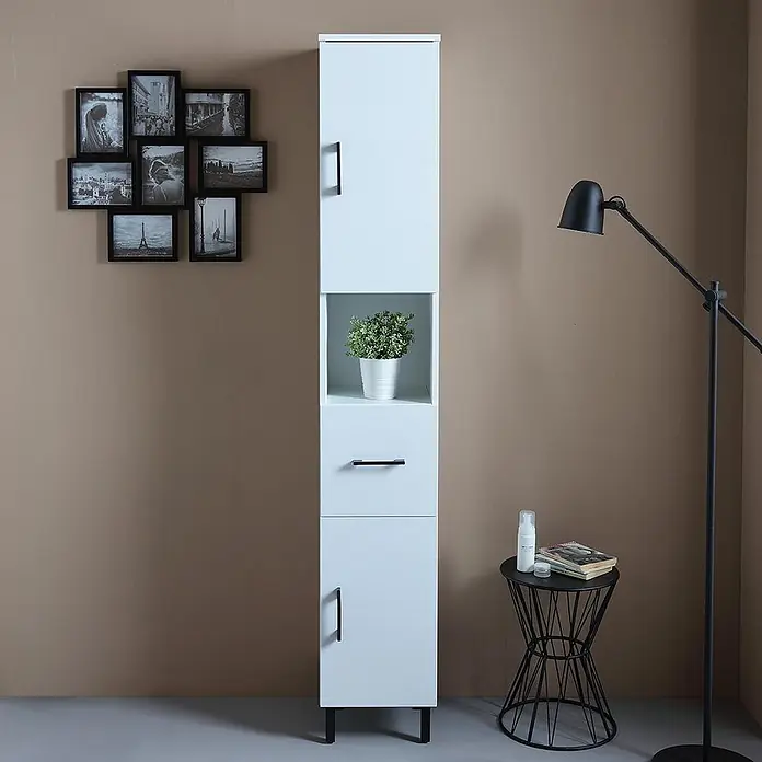 KV Badezimmerschrank hoch, weiß Export, reversibel mit Türen, Schublade und -  - Hoher weißer Stauraumschrank mit schwarzen Griffen, offenem Fach mit Pflanze, einer Schublade und zwei geschlossenen Fächern, neben einer Lampe und einem Beistelltisch.
