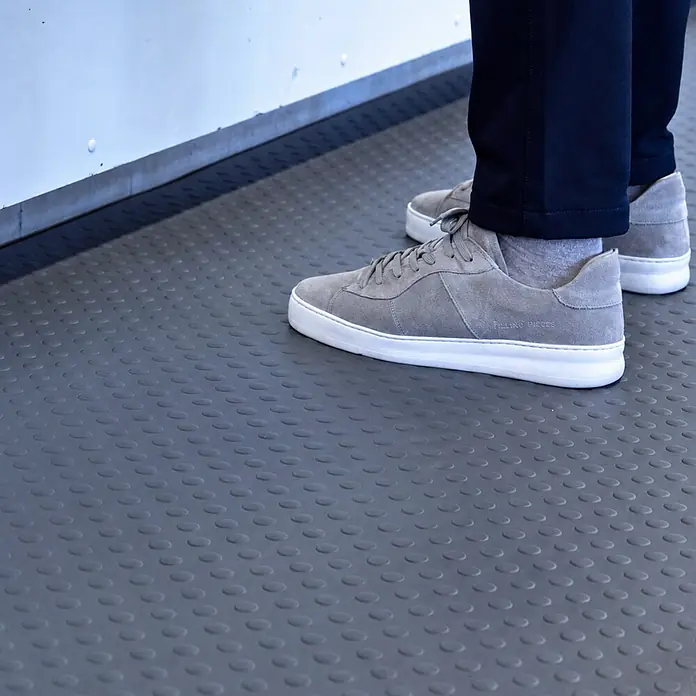 PROREGAL Anti-Ermüdungsmatte COMFORT PLUS mit Noppenprofil Grau -  - Eine Person trägt graue Wildleder-Sneaker mit weißen Sohlen, die auf einer dunkelgrauen strukturierten Matte stehen.