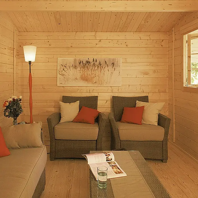 Kiehnholz Gartenhaus Östersund - Außenmaß inkl. Dachüberstand (B x T): 437 x 423 cm, Holz, Natur - Einladender Wohnbereich mit Holzvertäfelung, Korbsesseln und einem Couchtisch mit Zeitschrift und Glas Wasser. Gemütliche Atmosphäre mit Lampe und Kunstwerk.