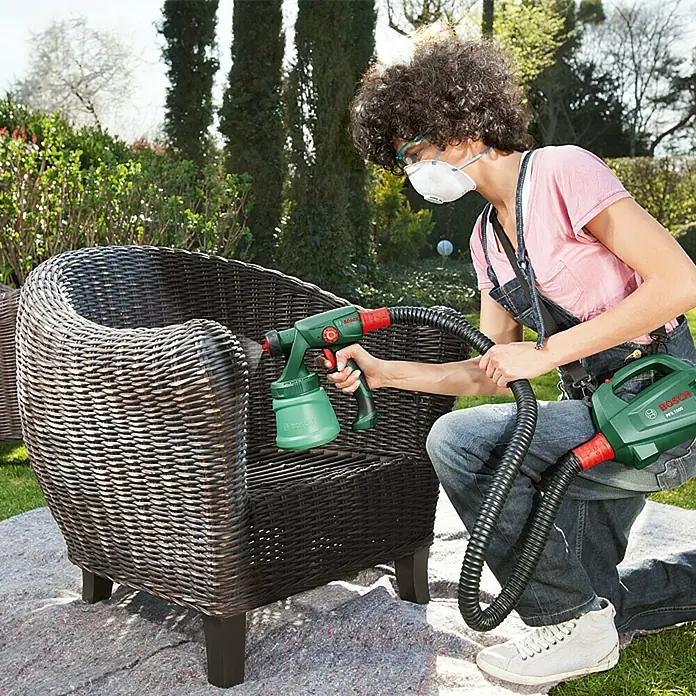 Bosch Sistema de pulverización de pintura PFS 1000 - Apto para: Esmaltes, Absorción nominal de corriente: 410 W, 800 ml - Una persona está pintando con aerosol una silla de ratán al aire libre con un pulverizador de pintura Bosch verde y rojo.
