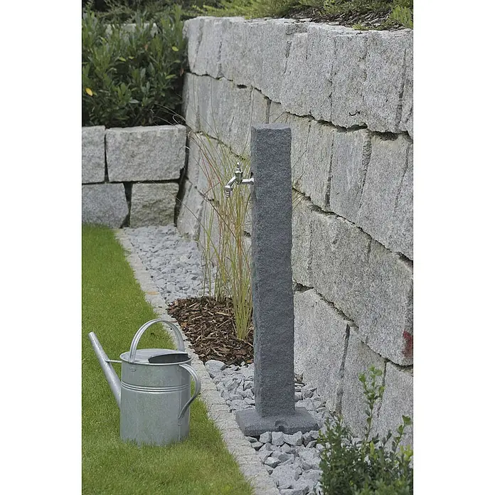 Arves Wasserzapfstelle Natura - Black Granitfarben, Höhe: 98 cm - Gartenszene mit Wasserhahn-Säule aus dunklem Stein, einer Zinkgießkanne und einer grauen Steinmauer im Hintergrund.