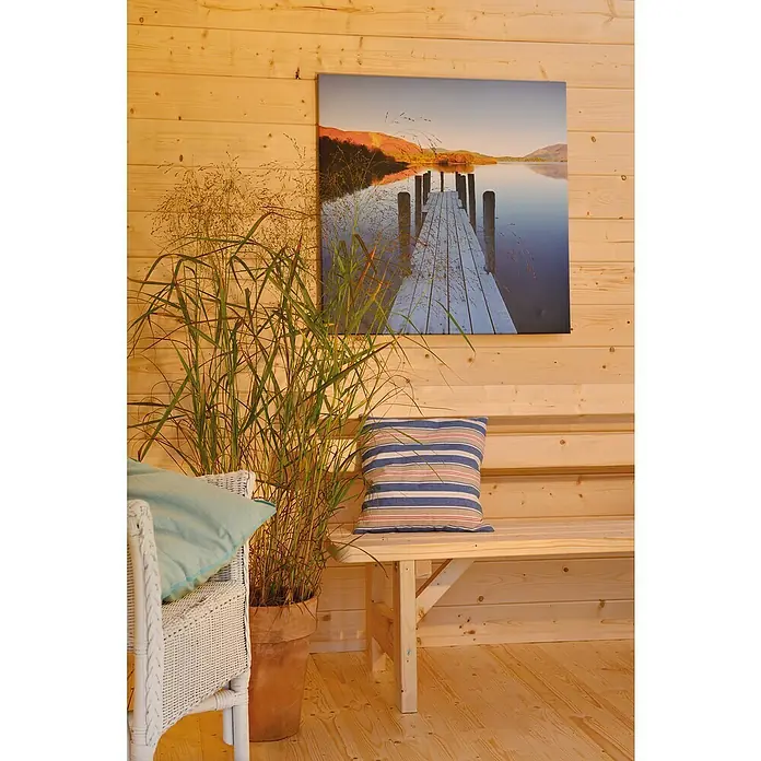 Kiehnholz Gartenhaus Stockholm 3 - Außenmaß inkl. Dachüberstand (B x T): 340 x 393 cm, Holz, Natur - Das Foto zeigt eine Zimmerpflanze in einem Topf, die neben einer Bank steht, die mit einem Kissen und einem weißen Rattansessel mit einem hellblauen Kissen dekoriert ist.