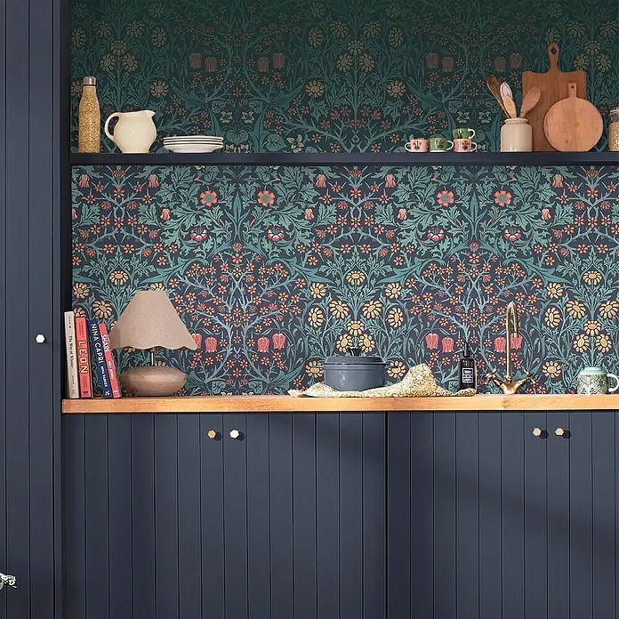 William Morris At Home Fototapete Blackthorn - Blau - Ein gemütlicher Küchenbereich mit dunkler floraler Tapete, Holzregalen und dunkelblauen Schränken, dekoriert mit Küchenutensilien und Büchern.