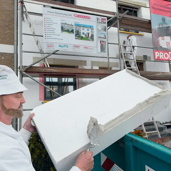 WDVS-Dämmplatte - Stärke: 100 mm, Wärmeleitstufe: 035, Expandiertes Polystyrol (EPS), Weiß - Ein Arbeiter trägt Dämmplatten auf eine Baustelle auf, mit einer "Haus-Sanierung mit BAUHAUS" Werbung im Hintergrund.