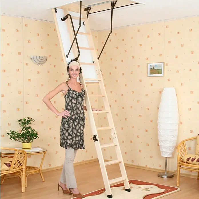 Star Stairs Bodentreppe Star Plus - 110 x 60 cm, Wärmedämmung: 0,85 W/(m²K) - Eine hölzerne Dachbodentreppe ist in einem Raum mit einer Frau zu sehen, die daneben steht. Die Treppe ist hell und robust.