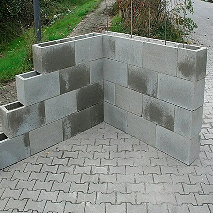 EHL Schalungsstein - 50 x 17,5 x 25 cm - Betonsteinmauer auf Pflastersteinen. Die grauen Betonsteine bilden eine Ecke und sind teilweise hohl.