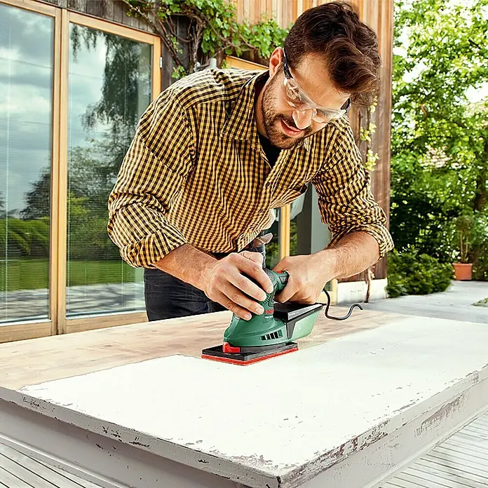 Bosch Elektro-Multischleifer PSM 200 AES - 200 W, Leerlaufschwingzahl: 6.000 U/min - 26.000 U/min, Durchmesser Schwingkreis: 1,8 mm - Mann schleift eine Oberfläche mit einem grünen Schleifgerät von Bosch. Das Gerät ist kompakt und handlich für präzises Arbeiten.