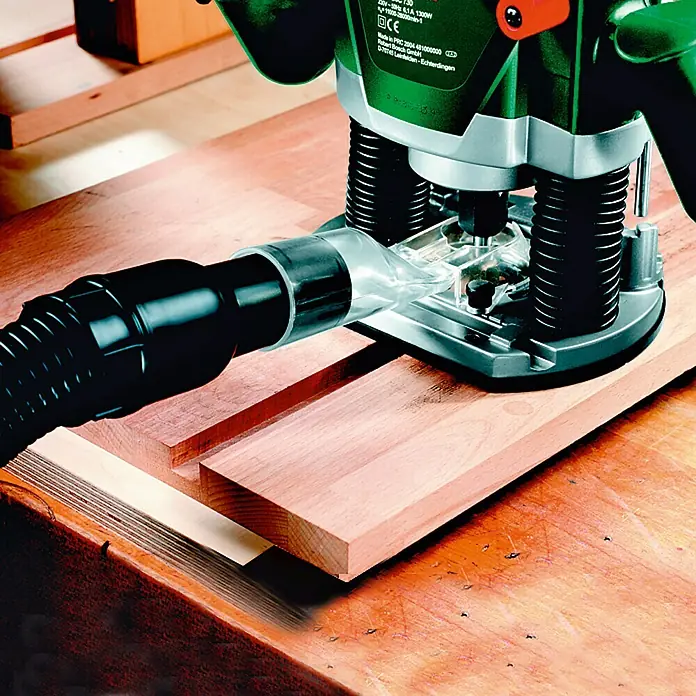 Bosch Fresadora eléctrica POF 1400 ACE - 1.400 W, 11.000 r.p.m. - 28.000 r.p.m., Diámetro del portaherramientas: 6 mm - 8 mm - Primer plano de una fresadora de madera con un accesorio de aspiración trabajando en una pieza de madera.