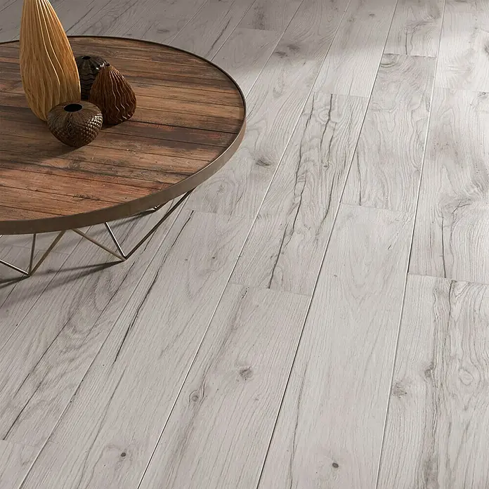 Pavimento porcelánico Genuine - 23,3 x 120 cm, Gris, Mate - Primer plano de un suelo de parqué con una mesa de centro rústica y jarrones decorativos. La madera clara crea un ambiente moderno y acogedor.