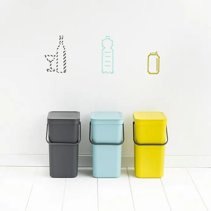 Brabantia Cubo de basura empotrado Sort & Go - 16 l, Gris, Plástico - Tres contenedores de reciclaje de colores (gris, azul, amarillo) con tapa y asas frente a una pared blanca con iconos de vidrio, plástico y latas.