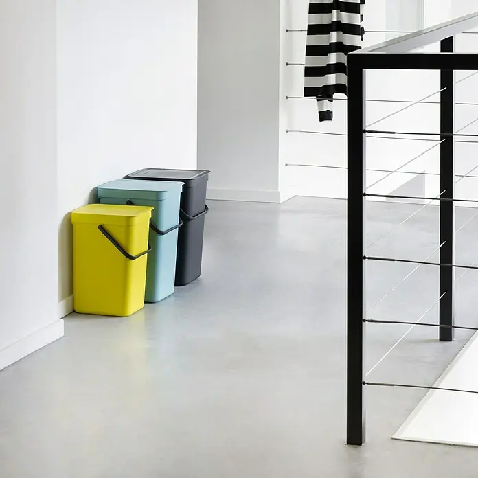 Brabantia Cubo de basura empotrado Sort & Go - 16 l, Gris, Plástico - Tres contenedores de basura de colores (amarillo, azul claro, gris oscuro) están contra una pared blanca, mientras que una barandilla moderna está en primer plano.