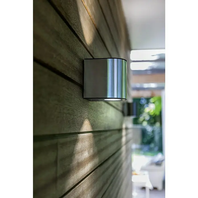 Lutec Aplique exterior LED Gemini - 24 W, 13,5 x 14,1 x 10,6 cm, Acero inoxidable, IP54 - Una lámpara de pared exterior moderna de color plateado ilumina un panel de madera, proporcionando una iluminación indirecta agradable. Diseño elegante.