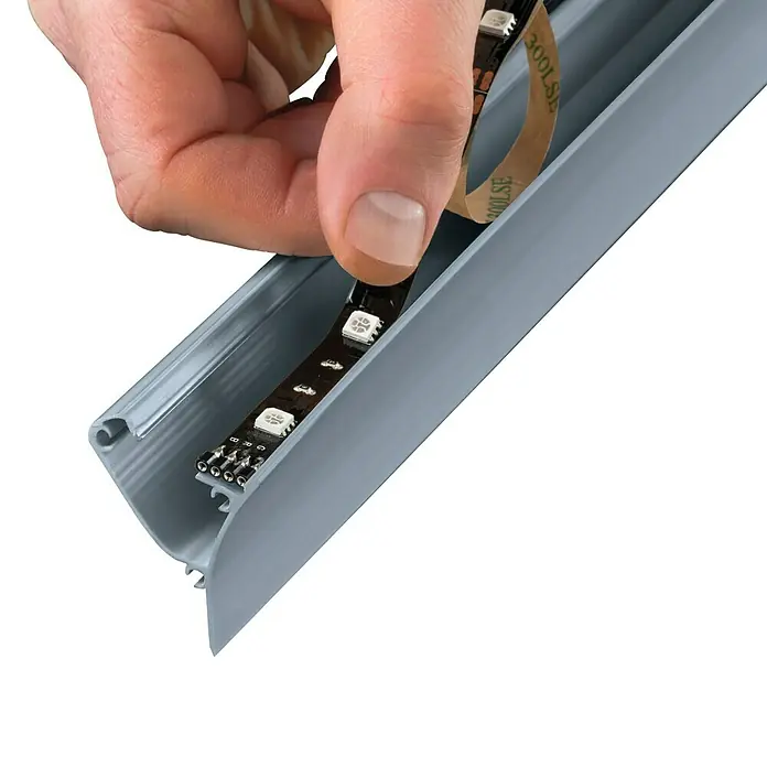 Paulmann Corner Profil - 1 m, Kunststoff - Nahaufnahme einer Hand, die ein LED-Lichtband mit Klebestreifen in ein graues Aluminiumprofil einsetzt. Ideal für indirekte Beleuchtung.