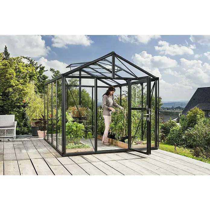Vitavia Gewächshaus Zeus 8100 - Außenmaß inkl. Dachüberstand (B x T): 256 x 316 cm, 3 mm, Schwarz - Ein Gewächshaus aus Glas mit einem offenen Eingang auf einer hölzernen Terrasse, in dem eine Frau Pflanzen pflegt.
