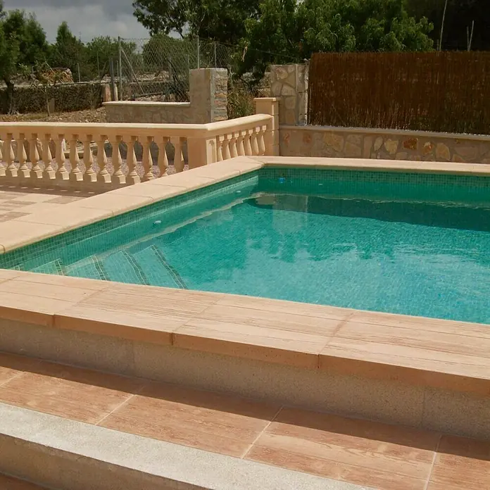 Pavimento Remate piscina Teka - 50 x 45 cm, Beige - Primer plano de una piscina con azulejos celeste y escalones de piedra, bordeada por una balaustrada y vegetación exuberante al fondo.