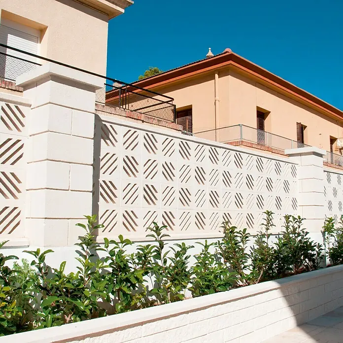 Bloque Pilastra Liso 2 - 30 x 30 x 20 cm, Blanco, Hormigón, Liso - Toma de una pared blanca con un patrón diagonal cuadrado, que se extiende frente a edificios y bajo un cielo azul brillante.