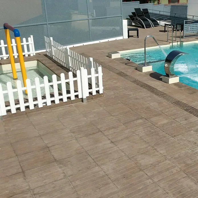 Pavimento para exterior Madera Nogal - 50 x 50 cm, Marrón - Vista de una zona de piscina con una fuente de agua y una piscina para niños, rodeada por una valla blanca.