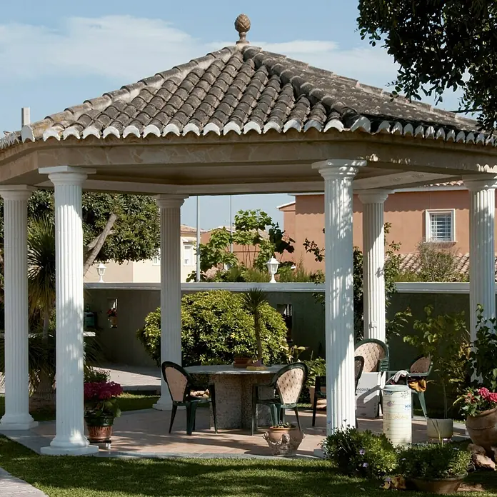 Base para techo o suelo Capitel Atenas - 40 x 40 cm - Un mirador con columnas y una mesa de piedra con sillas en un jardín, rodeado de plantas. El mirador ofrece un espacio sombreado para la relajación.