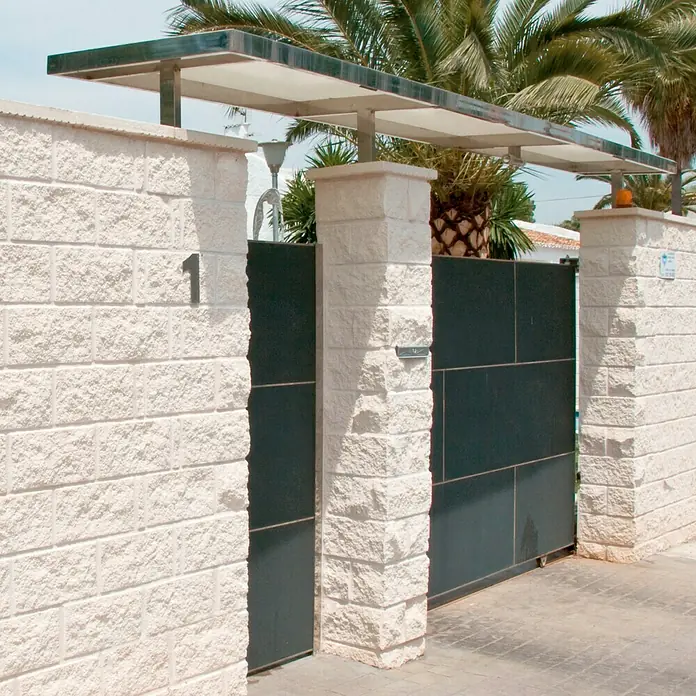 Bloque Pilastra Split - 27 x 27 x 20 cm, Blanco, Hormigón, Ligeramente irregular - Puerta cerrada con pilares de piedra y número de casa. Las palmeras bordean la entrada, proporcionando privacidad y sombra.