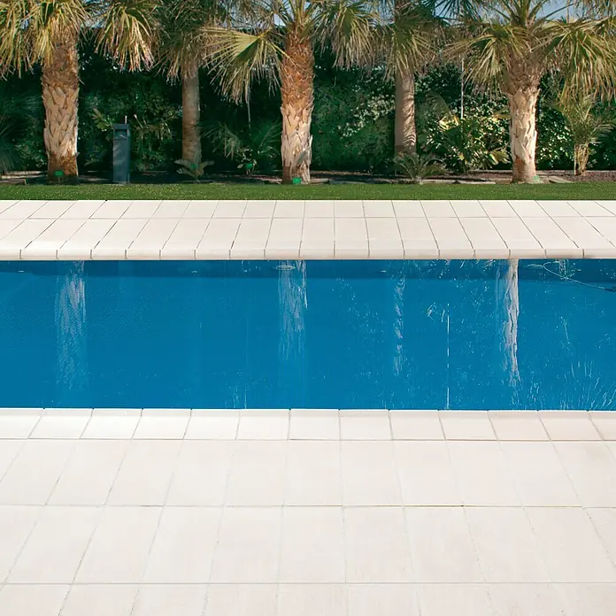 Pavimento remate Piscina R1 - 50 x 25 cm, Blanco - Vista de una piscina rectangular con azulejos blancos y palmeras al fondo, invitando a la relajación y el disfrute.