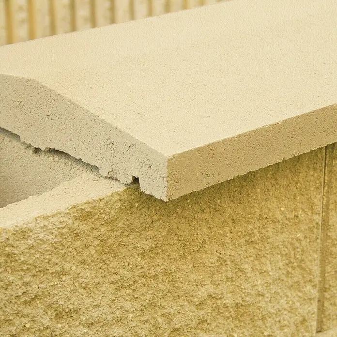 Cubremuros doble pendiente - 50 x 27 x 5 cm, Crema, Hormigón - Primer plano de una valla de bloques de hormigón beige con una tapa lisa, que contrasta con la textura rugosa de la piedra.\n