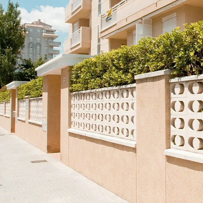 Celosía de pared Mundo - 40 x 20 cm, Blanco - Un elegante muro de hormigón con aberturas decorativas y vegetación exuberante junto a una acera soleada.