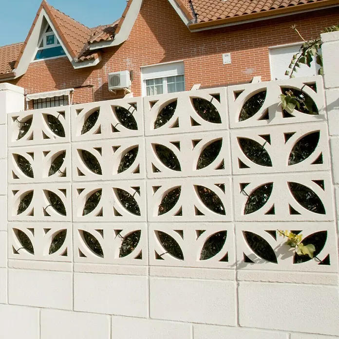 Celosía de pared Pétalos - 40 x 20 cm, Blanco - Una valla blanca decorativa con un patrón de hojas frente a una casa de ladrillo rojo bajo un cielo azul ofrece privacidad y atractivo estético.