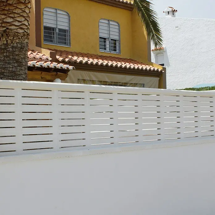 Celosía de pared Cebra - 40 x 20 cm, Blanco - Primer plano de una valla blanca frente a una casa amarilla con tejas rojas. La valla proporciona privacidad y seguridad a la propiedad.