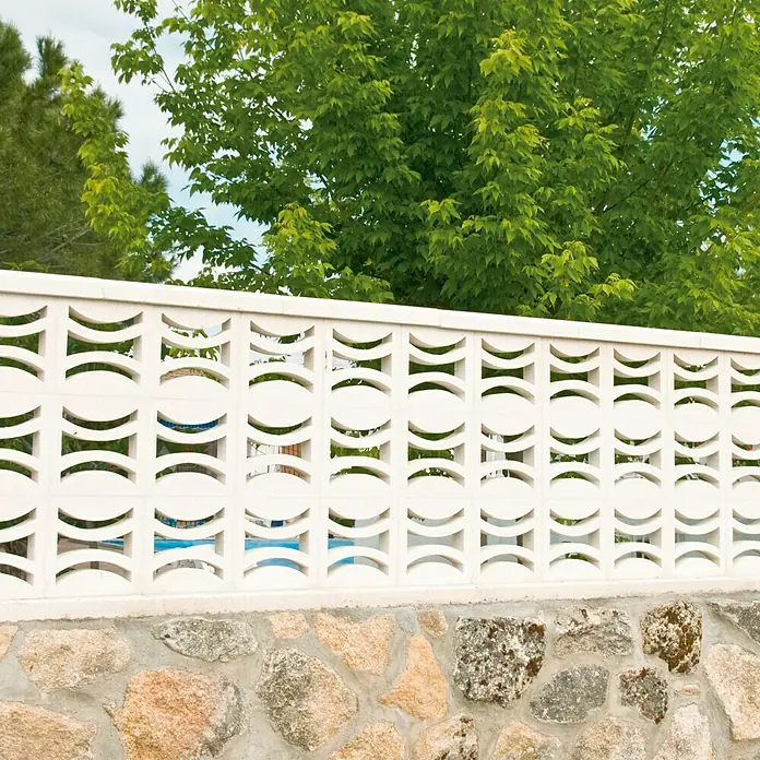 Celosía de pared Flor - 25 x 25 cm, Blanco - Primer plano de una valla blanca decorativa con base de piedra, rodeada de exuberante vegetación, formando un elegante límite exterior.