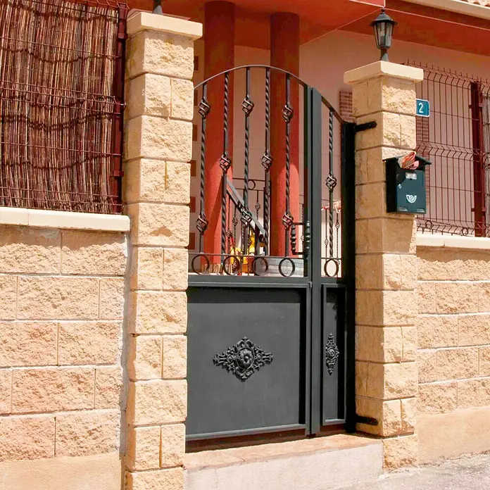 Bloque Pilastra Split - 40 x 40 x 20 cm, Crema, Hormigón, Ligeramente irregular - Vista de una puerta de metal negro adornada que se encuentra entre pilares de piedra, con un buzón y un número de casa.
