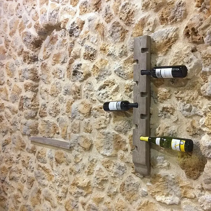 Estantería para botellas Nogal - 90 x 10 x 5,5 cm - Un soporte de madera para botellero con tres botellas de vino fijadas a una pared de piedra.